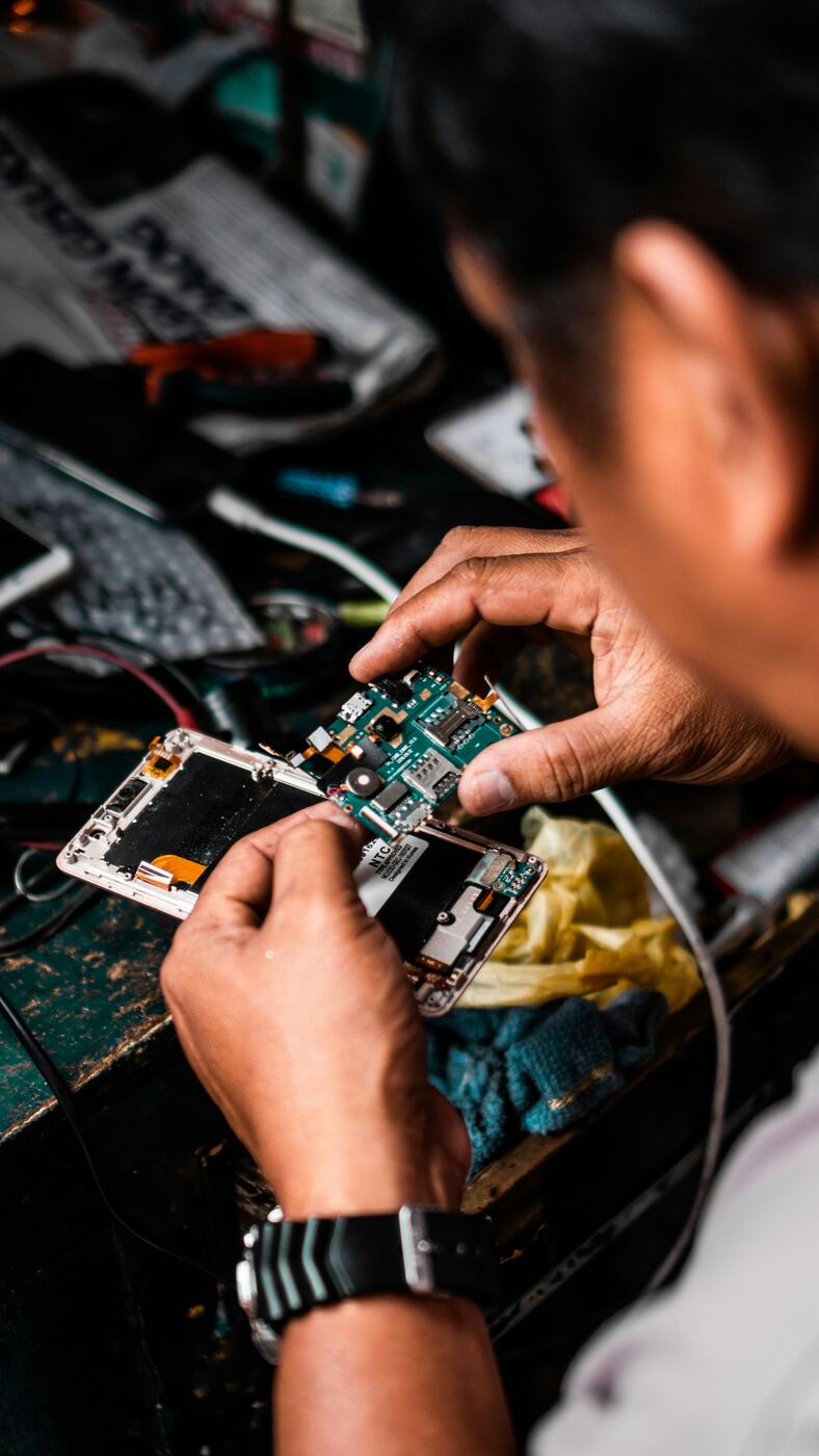 Hands at a workshop bench repairing an opened Android smartphone — close focus on the circuit board, with cables and tools in the background.